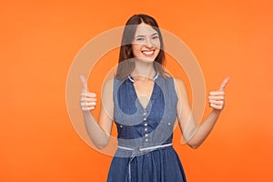 Like, approval gesture. Delighted happy brunette woman in denim dress smiling broadly and giving double thumbs up