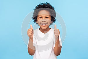 Like! Portrait of happy little boy with curly hair in white T-shirt smiling at camera and doing thumbs up gesture