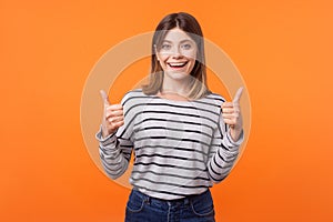 Like, thumbs up! Portrait of joyful kind young woman with brown hair in long sleeve striped shirt. isolated on orange background