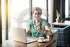 Like! Young happy businessman in green t-shirt sitting and working on laptop, looking at camera with thumbs up and toothy smile