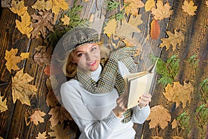 She likes detective genre. Girl blonde lay wooden background with leaves. Woman lady in checkered hat and scarf read
