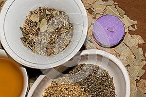 A lilac candle extinguished next to three white bowls with medicinal herbs and tea on dried leaves