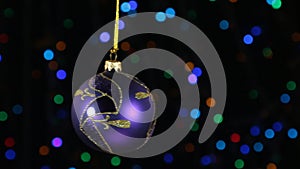 Lilac Christmas ball swinging on defocused background with flashing garland lights.