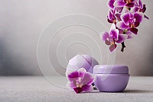Lilac cosmetic cream jar and bath ball on gray background with orchid flower