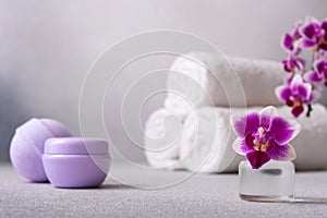 Lilac cosmetic cream jar, bath ball and rolled up towels on gray background with orchid flower