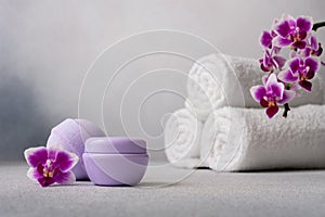 Lilac cosmetic cream jar, bath ball and rolled up towels on gray background with orchid flower