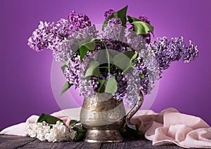 Lilac flowers in an old vase, near sprig of white flowers on background of fine fabric