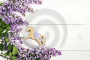 Lilac flowers with toy rocking horse on white wooden background.