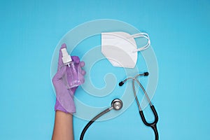 Lilac medical gloved hand holding disinfectant spray, white surgical mask and stethoscope on light blue background