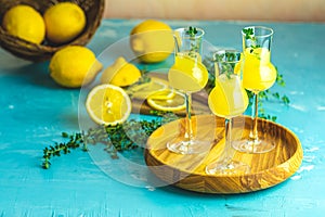 Limoncello with thyme in three grappas wineglass in wooden tray, fresh lemon in basket on light concrete table. Artistic still photo