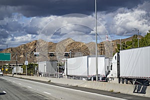 Line of big rig semi trucks with loaded semi trailers standing on the at the weighing station with scales for weighing along the