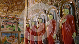 Line of Buddhist statues in ornate temple. Golden walls, intricate murals scenes. Robed figures stand serene, symbols of