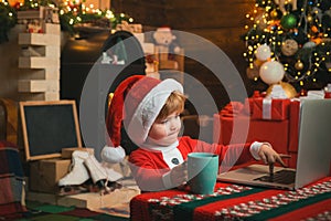 On-line Christmas shopping for kids. Little kid is wearing Santa clothes sitting by his laptop. Santa helper using