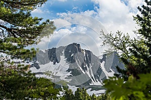 Line of mountains on horizon under summer sky. Coniferous forest on hillside, rocks are covered with snow