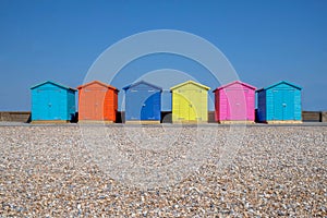 A line of six multicoloured beach huts on Seaford beach, in the foreground is a pebble beach in the back ground is a clear blue