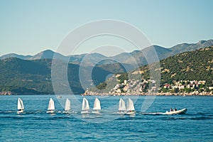 A line of small sailing boats with kid sailors on a tugboat is passing by the hilly seashore