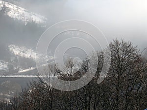 Linear traces of vanishing roads into the fog, among the steep mountains with bare trees in the foreground