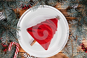 Linen napkin folded in shape of Christmas tree on white plate among spruce branches. New Year table setting. Top view