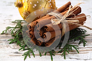 Linking of sticks of cinnamon on fir-tree branches, close up on a wooden light background, with a Christmas tree decoration on a b