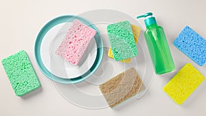 Liquid dishwashing detergent with heap of colorful sponge and dishes on light beige background