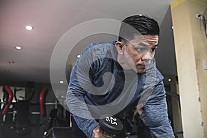A listless and seemingly unmotivated asian man makes dumbbell triceps kickbacks on a bench at a gym. Tricep and arm exercise and