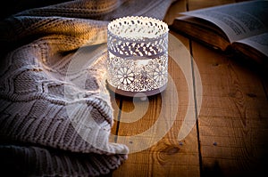 Lit candle in a lace candle holder, knitted sweater and an open book on wood background, cozy atmosphere
