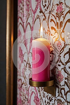 A lit candle rests on a gold-toned wall mount, casting a warm glow against vibrant pink and white floral photo