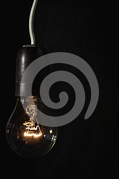 Lit incandescent lamp with tungsten filament glowing on the black background with copy space for text. Saving energy concept.