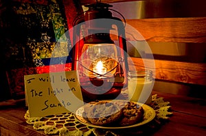 Lit lamp sitting next to a plate of cookies at night and a note for Santa.