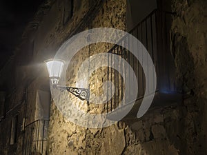 A lit streetlamp illuminates an aged wall and balcony at night, creating a moody, atmospheric, and historical scene