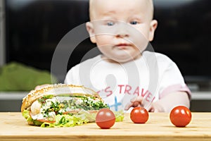 Litlle infant caucasian boy is trying to steal one unhealthy delicious hotdog with green salad from the table