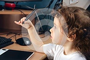 Little 2 3 year old baby girl in white clothers draws at the home computer in graphics drawing tablet. The child is holding a pen