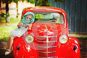 Little 2 years old girl in retro style with old fashioned camera in hands, white dress, sunglasses and green scarf on head sit on