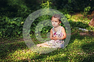 Little 3 years old short haired girl in gray dress sits on green grass in garden with small pink elephant toy. Sad child in summer