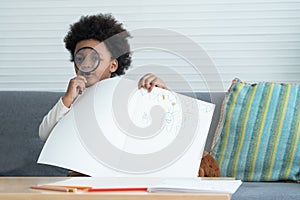 Little active African kids boy playing showing his drawing on book, holding magnifying glass and sitting on sofa at home