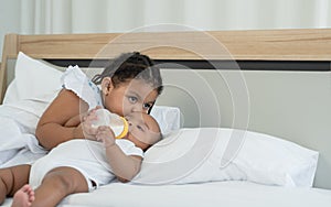 Little adorable African older sister feeding newborn baby milk bottle and kissing with love and care while infant lying on bed