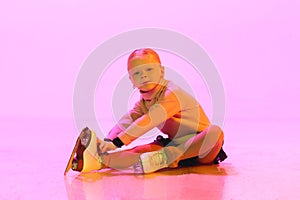 Little adorable baby girl, chide in sweater and skirt, sitting on floor and tying laces on skates against pink