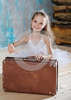 Little adorable ballerina in white tutu with old vintage suitcase in a beautiful studio. Travel and voyage