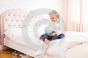 Little adorable blonde girl in school uniform and white golfs siting on a ber in her bedroom with globe in her hands photo