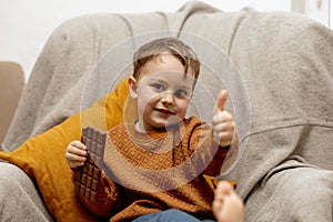 Little adorable boy sitting on the couch at home and eating chocolate bar. Child and sweets, sugar confectionery. Kid