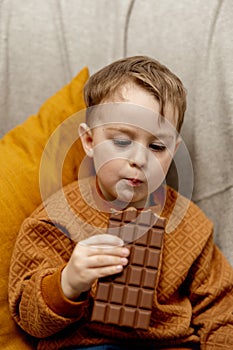 Little adorable boy sitting on the couch at home and eating chocolate bar. Child and sweets, sugar confectionery. Kid