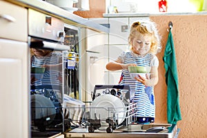 Little adorable cute toddler girl helping to unload dishwasher. Funny happy child standing in the kitchen, holding