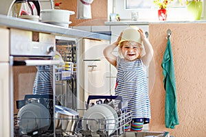 Little adorable cute toddler girl helping to unload dishwasher. Funny happy child standing in the kitchen, holding
