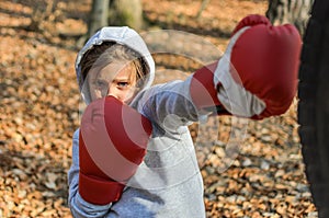 Little adorable girl child, in a sports uniform in a hood, trains in boxing, in the forest, in red boxing gloves hits a pear from