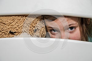 Little adorable girl child with a teddy bear looks through a crack in a hole in a wooden fence