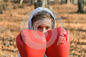 Little adorable girl child in a tracksuit, with a hood on his head, in red boxing gloves, boxing on the street in the autumn fores