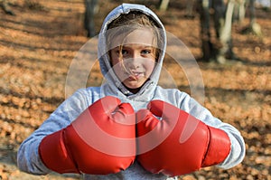 Little adorable girl child in a tracksuit, with a hood on his head, in red boxing gloves, boxing on the street in the autumn fores