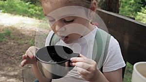 Little adorable girl with long black eyelashes and white T-shirt blows on hot tea in mug from thermos during forest hike