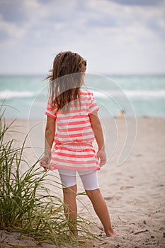 Little adorable kid girl in striped dress standing on the sandy beach and looking at the ocean. Sea vacations. Child dreaming.
