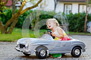 Little adorable toddler girl driving big vintage toy car and having fun with playing outdoors. Crying sad upset child on